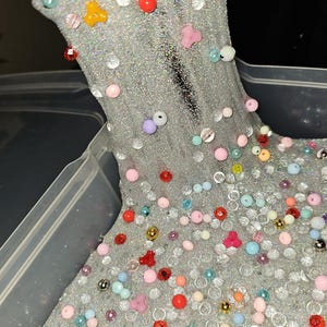 Peut inclure: Slime argenté scintillant avec des perles et des embellissements colorés. Le slime est rempli de petites perles rondes transparentes et de perles colorées de différentes tailles, dont des rouges, roses, jaunes et bleues. Le slime est dans un récipient en plastique transparent.