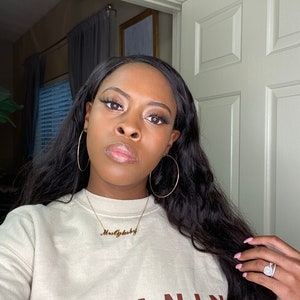 May include: A woman wearing a beige sweatshirt with the word "MELANIN" in brown block letters. She is wearing gold hoop earrings and a gold necklace that says "MrsOglsby". Her long, dark hair is wavy.