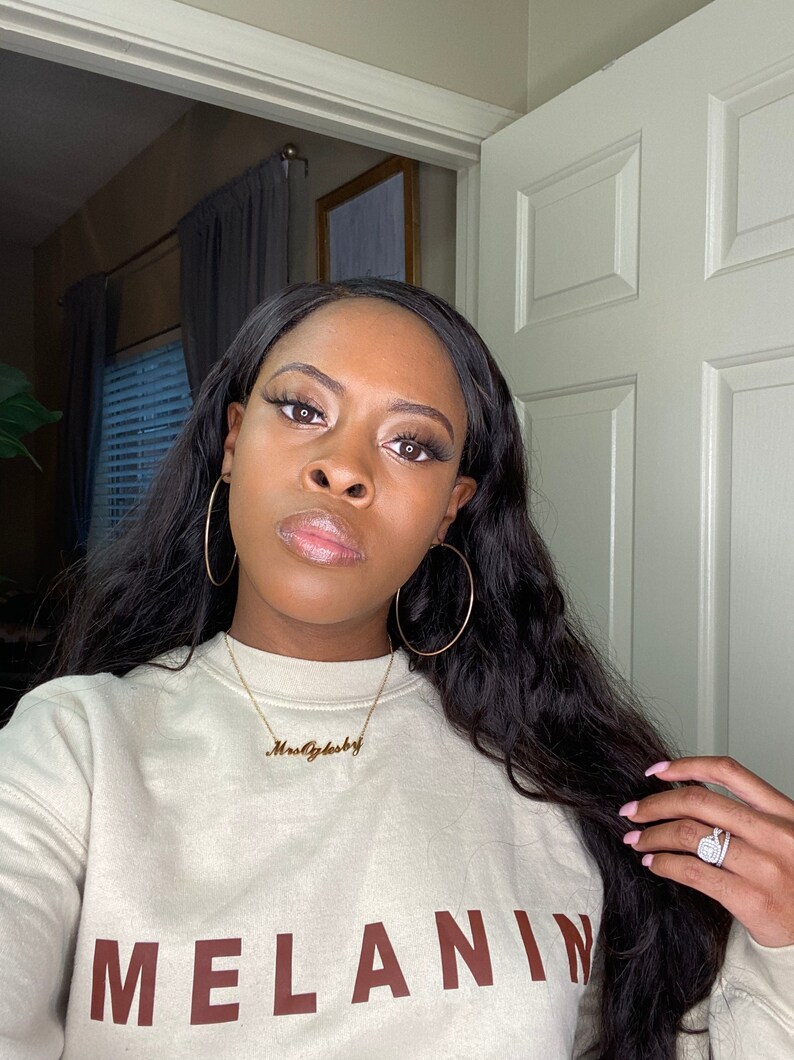 May include: A woman wearing a beige sweatshirt with the word "MELANIN" in brown block letters. She is wearing gold hoop earrings and a gold necklace that says "MrsOglsby". Her long, dark hair is wavy.