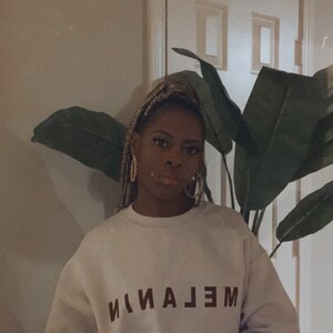 May include: A light-colored sweatshirt with the word "MELANIN" printed in dark brown. The sweatshirt has long sleeves and is worn by a person. The background includes a large-leafed plant and a white door.