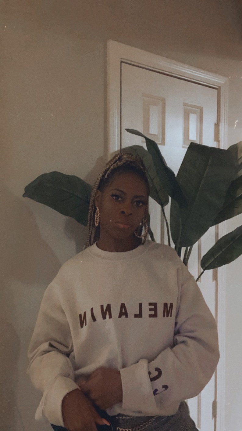 May include: A light-colored sweatshirt with the word "MELANIN" printed in dark brown. The sweatshirt has long sleeves and is worn by a person. The background includes a large-leafed plant and a white door.
