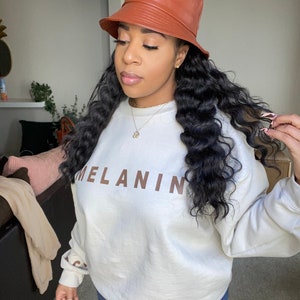 May include: A brown leather bucket hat and a cream-colored sweatshirt with the word "MELANIN" printed in brown. The model is wearing blue jeans.