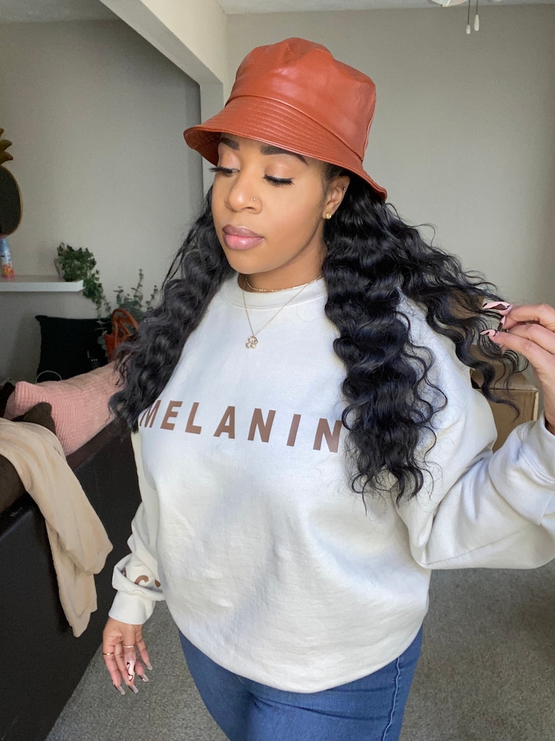 May include: A brown leather bucket hat and a cream-colored sweatshirt with the word "MELANIN" printed in brown. The model is wearing blue jeans.