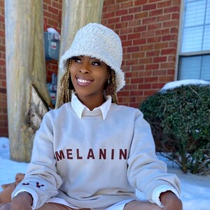May include: A cream-colored, fuzzy bucket hat paired with a light beige sweatshirt. The sweatshirt features the word "MELANIN" in maroon lettering across the chest. The sleeves have a small embroidered design. The person is wearing a white collared shirt underneath.