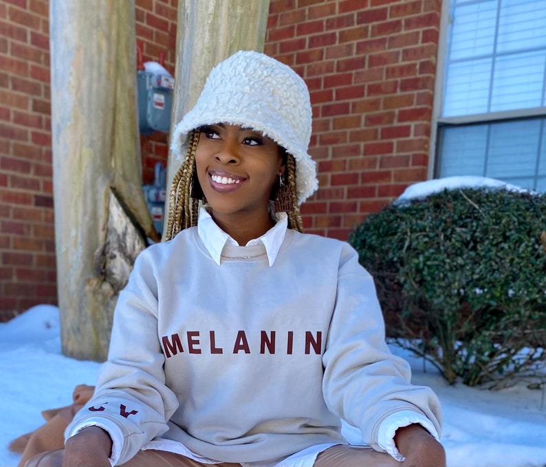 May include: A cream-colored, fuzzy bucket hat paired with a light beige sweatshirt. The sweatshirt features the word "MELANIN" in maroon lettering across the chest. The sleeves have a small embroidered design. The person is wearing a white collared shirt underneath.