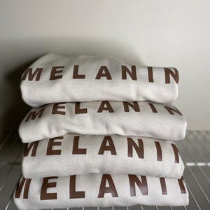 May include: A stack of four folded cream-colored sweatshirts. Each sweatshirt has the word "MELANIN" printed in a dark brown, bold font across the front. The sweatshirts are neatly arranged on a white wire shelf.