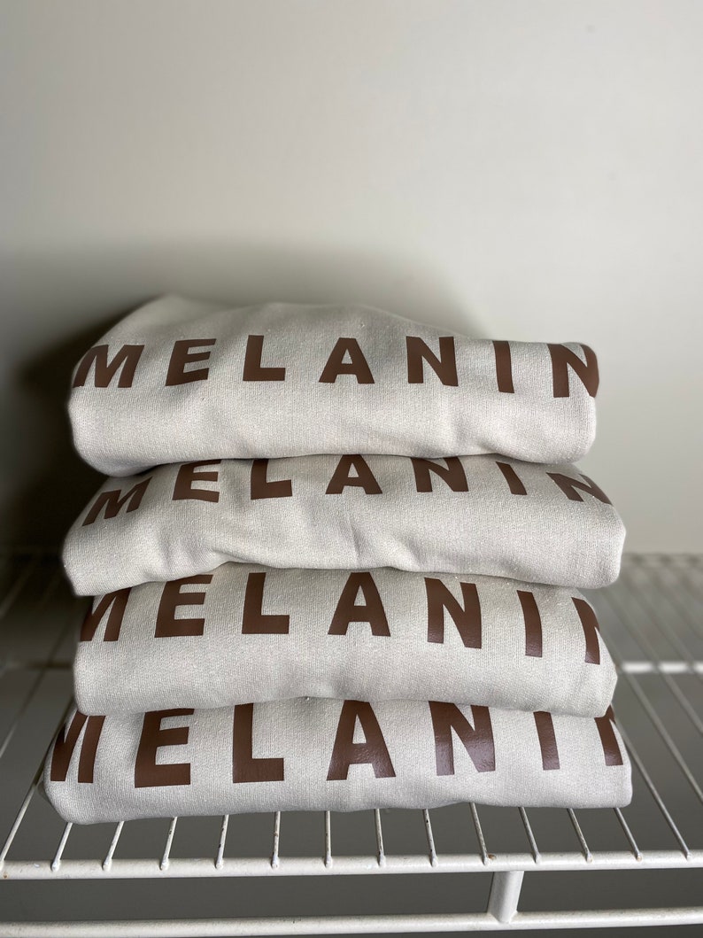 May include: A stack of four folded cream-colored sweatshirts. Each sweatshirt has the word "MELANIN" printed in a dark brown, bold font across the front. The sweatshirts are neatly arranged on a white wire shelf.