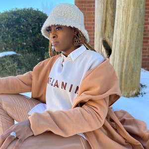 May include: A woman wearing a cream-colored, fuzzy bucket hat and hoop earrings with the text "Black Lives Matter." She is wearing a white and brown sweatshirt with the word "MELANIN" printed on it, a tan coat, and tan pants.