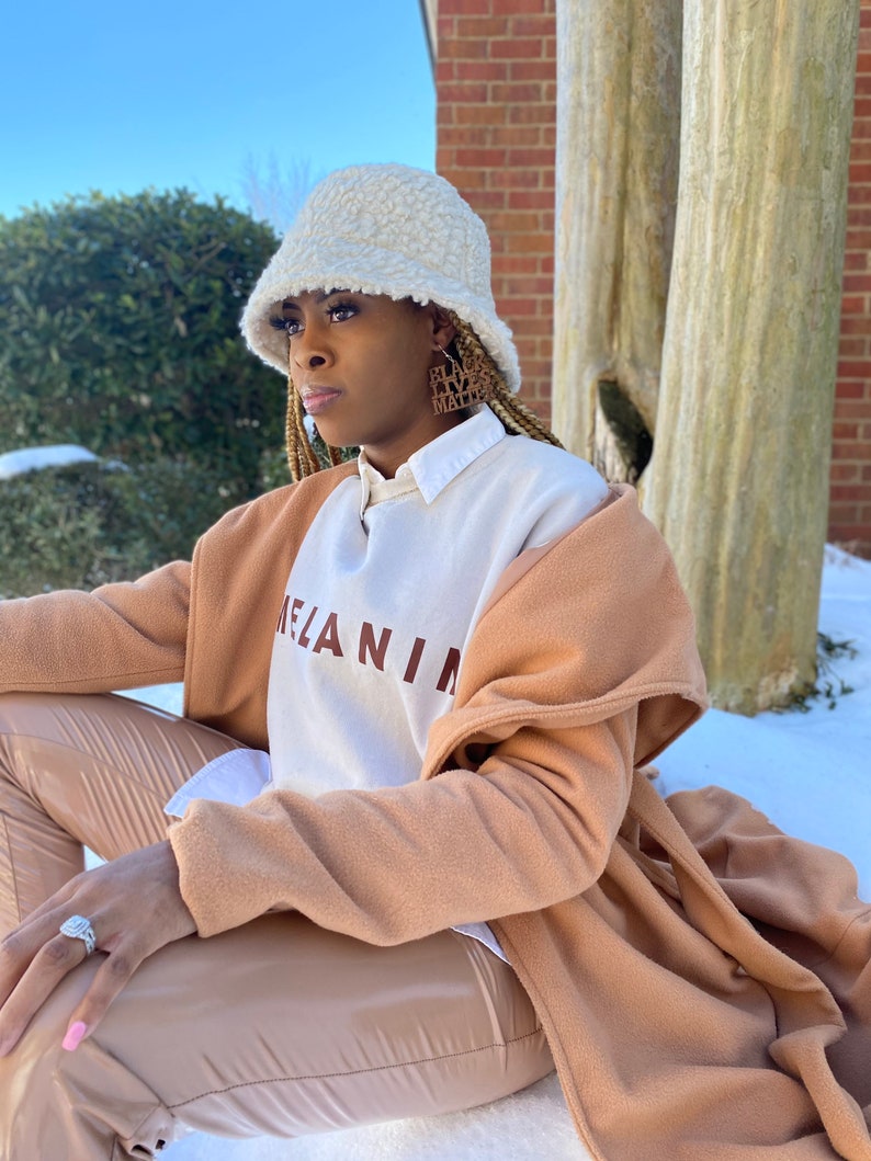 May include: A woman wearing a cream-colored, fuzzy bucket hat and hoop earrings with the text "Black Lives Matter." She is wearing a white and brown sweatshirt with the word "MELANIN" printed on it, a tan coat, and tan pants.