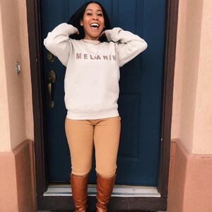 May include: A person wearing a cream-colored sweatshirt with the word "MELANIN" printed in brown, paired with tan pants and brown knee-high boots. The person is standing in front of a blue door.
