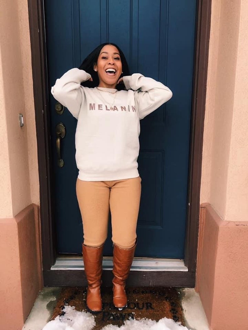 May include: A person wearing a cream-colored sweatshirt with the word "MELANIN" printed in brown, paired with tan pants and brown knee-high boots. The person is standing in front of a blue door.