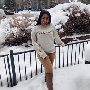 May include: A beige sweatshirt with the word "MELANIN" in brown letters. The sweatshirt has long sleeves and is paired with brown pants and brown knee-high boots. The background features a snowy landscape and a black metal fence.