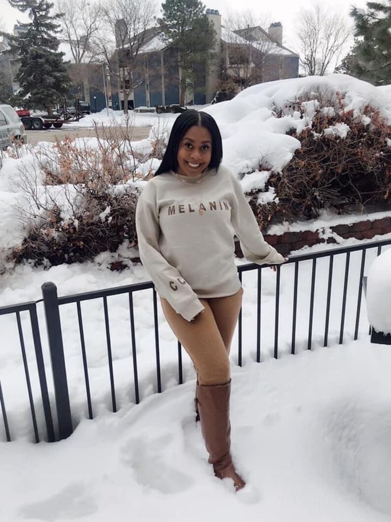 May include: A beige sweatshirt with the word "MELANIN" in brown letters. The sweatshirt has long sleeves and is paired with brown pants and brown knee-high boots. The background features a snowy landscape and a black metal fence.