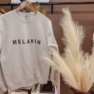 May include: A cream-colored sweatshirt with the word "MELANIN" printed in brown block letters. The sweatshirt hangs on a wooden hanger on a black clothing rack. A decorative vase with tall, feathery beige grass is in the foreground.