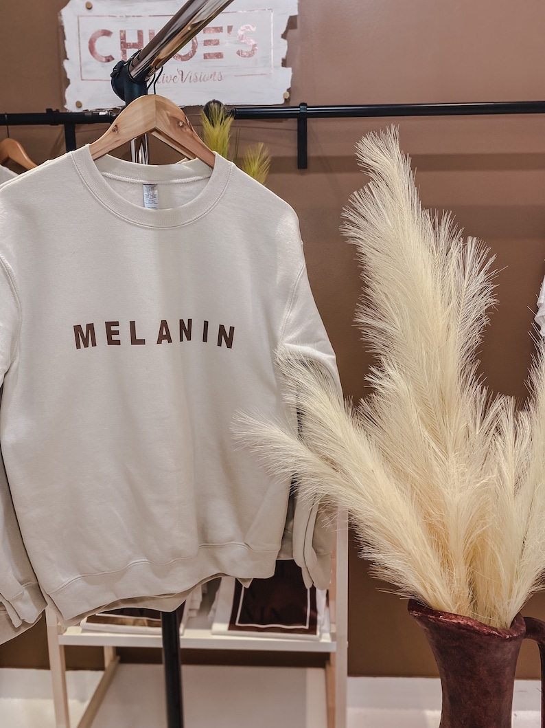 May include: A cream-colored sweatshirt with the word "MELANIN" printed in brown block letters. The sweatshirt hangs on a wooden hanger on a black clothing rack. A decorative vase with tall, feathery beige grass is in the foreground.