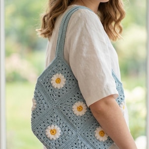 May include: A light blue crochet tote bag featuring a repeating pattern of white daisy motifs with yellow centres. The bag is constructed from square panels joined together, with two integrated shoulder straps made of the same blue yarn.