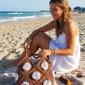 May include: A brown crochet tote bag constructed from square panels, each featuring a central white flower motif with a light yellow centre. The bag has two integrated handles and a diamond-patterned texture throughout the brown yarn body.