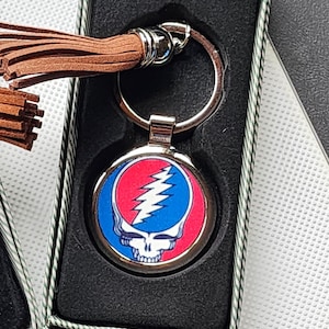 May include: A silver keychain with a round, blue, red, and white graphic of a skull with a lightning bolt. The keychain has a brown tassel.