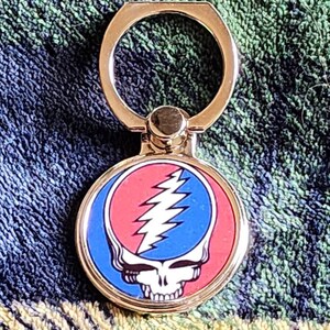 May include: A gold phone ring holder with a circular design featuring the Grateful Dead's iconic Steal Your Face logo in red, white, and blue.