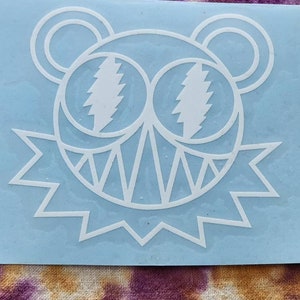 May include: White vinyl sticker of a cartoon bear with lightning bolt eyes and a jagged mouth. The bear is outlined in white and has a blue background.