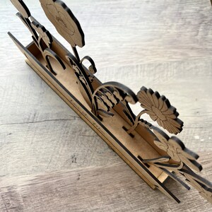 May include: Wooden flower stand with a cutout design of various flowers, including a poppy, lily of the valley, and a daisy. The stand has a brown wood finish and the text "Love is in the air" is written on the front.