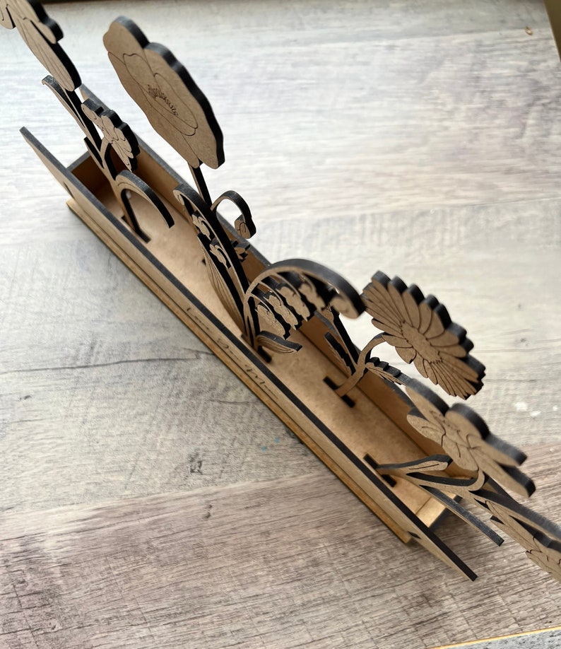 May include: Wooden flower stand with a cutout design of various flowers, including a poppy, lily of the valley, and a daisy. The stand has a brown wood finish and the text "Love is in the air" is written on the front.