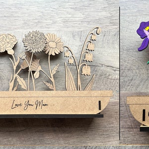 May include: Two wooden flower pot decorations. The first has a variety of flowers and the text "Love You Mom". The second has a purple iris and the text "Love Grows Here".