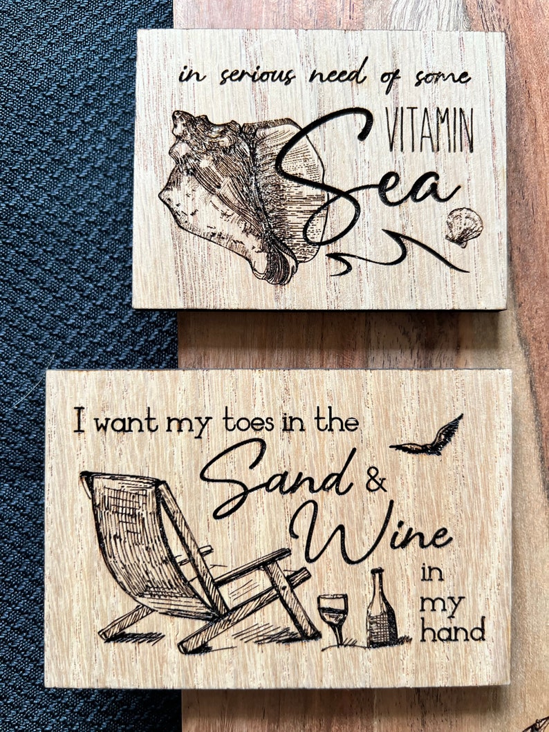 May include: Two wooden signs with a beach theme. The top sign has a seashell and the text "in serious need of some VITAMIN Sea". The bottom sign has a beach chair, a glass of wine, a bottle of wine, and the text "I want my toes in the Sand & Wine in my hand".