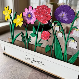 May include: A wooden craft with a white background and the text "Love You Mom". The craft features a variety of colourful flowers, including yellow daffodils, pink and purple cosmos, red roses, purple morning glories, and white lily of the valley.