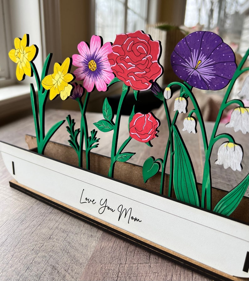 May include: A wooden craft with a white background and the text "Love You Mom". The craft features a variety of colourful flowers, including yellow daffodils, pink and purple cosmos, red roses, purple morning glories, and white lily of the valley.