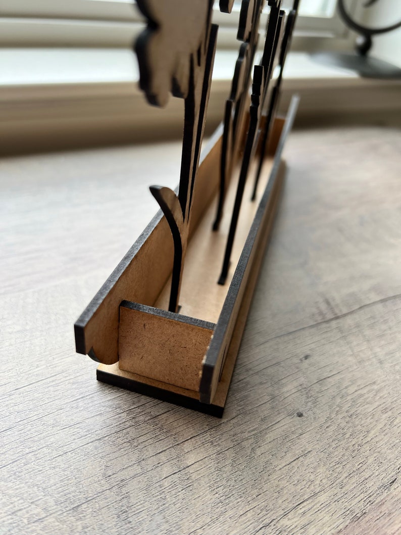 May include: A wooden flower stand with a black painted finish. The stand has a rectangular base with slots for holding flowers. The stand is made of plywood and has a rustic look.
