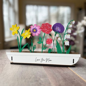 May include: A white wooden planter with a variety of colourful paper flowers, including yellow daffodils, pink cosmos, red carnations, purple morning glories, and white lily of the valley. The planter has the text "Love You Mom" written on the front.