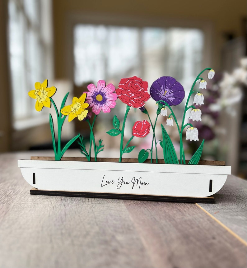 May include: A white wooden planter with a variety of colourful paper flowers, including yellow daffodils, pink cosmos, red carnations, purple morning glories, and white lily of the valley. The planter has the text "Love You Mom" written on the front.