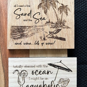 May include: Two wooden signs with a beach theme. The top sign has a drawing of palm trees and a beach with the text "all I need is the Sand Sea and the wine... lots of wine!" The bottom sign has a drawing of a beach umbrella, a cocktail, and flip flops with the text "totally obsessed with the ocean I might be an aquaholic".