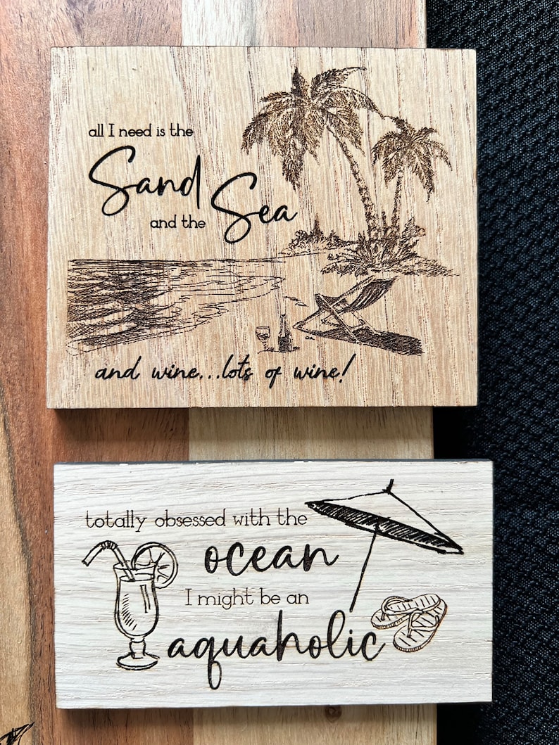 May include: Two wooden signs with a beach theme. The top sign has a drawing of palm trees and a beach with the text "all I need is the Sand Sea and the wine... lots of wine!" The bottom sign has a drawing of a beach umbrella, a cocktail, and flip flops with the text "totally obsessed with the ocean I might be an aquaholic".