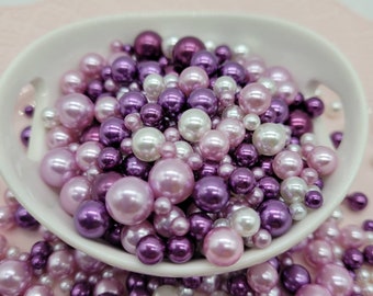 May include: A white bowl filled with a mixture of purple, pink, and white pearl beads. The beads are various sizes and have a glossy finish.