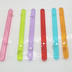 May include: Six colorful, glittery, plastic sticks. The sticks are pink, green, purple, red, blue, and orange.