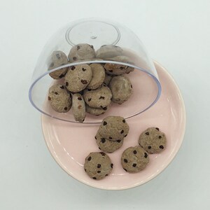 May include: A clear plastic container filled with chocolate chip cookies, with some cookies spilling out onto a pink plate.