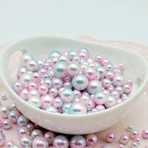 May include: A white bowl filled with iridescent pastel pink, blue, and white pearl beads. The beads are scattered around the bowl on a pink surface.
