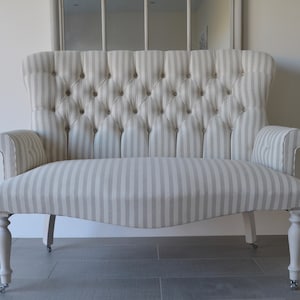May include: A small, white and grey striped sofa with a tufted back and wooden legs. The sofa has a curved shape and is upholstered in a linen fabric.