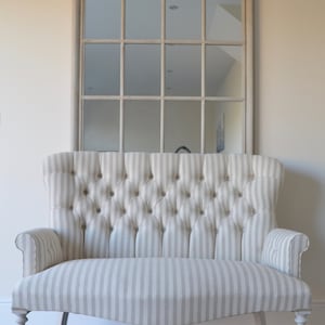 May include: A white and grey striped loveseat with a tufted back and rolled arms. The loveseat has a curved shape and sits on four turned legs with castors. The loveseat is in front of a large arched mirror with a white frame.