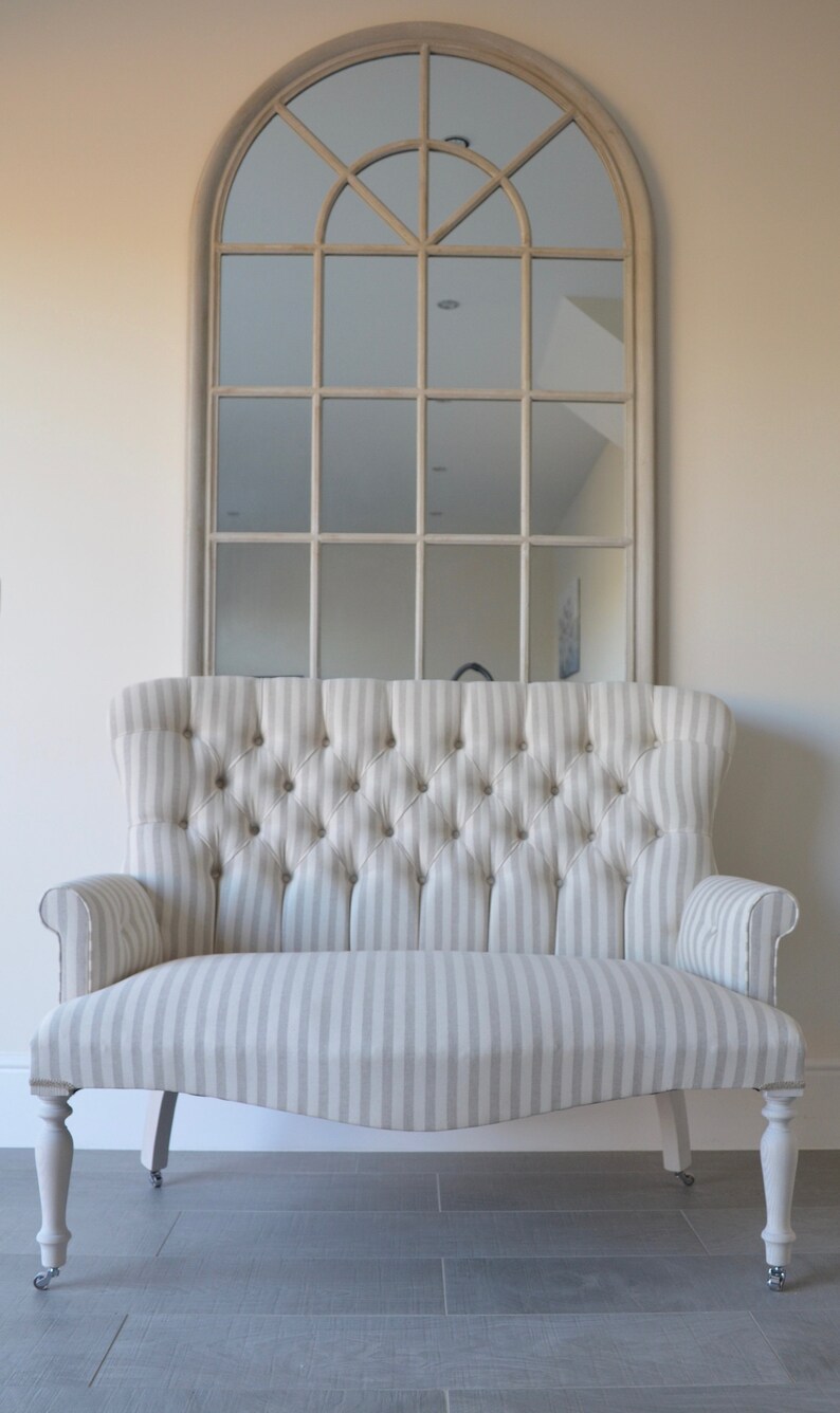 May include: A white and grey striped loveseat with a tufted back and rolled arms. The loveseat has a curved shape and sits on four turned legs with castors. The loveseat is in front of a large arched mirror with a white frame.