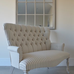 May include: A small, white and grey striped loveseat with button tufting and wooden legs. The loveseat is sitting on a grey tiled floor in front of a large arched mirror.