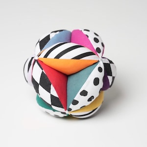 Op de afbeelding: Een kleurrijke stoffen babybal met verschillende geometrische patronen. De bal heeft panelen met zwart-witte strepen, stippen en dambordpatronen, samen met effen panelen in oranje, rood, teal en paars. Een zacht speelgoed.