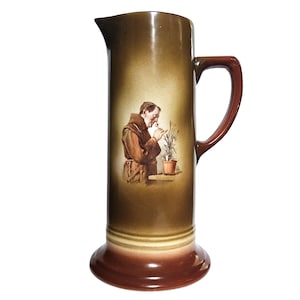May include: Tall, brown ceramic pitcher with a dark brown handle and base. The pitcher features a painted image of a monk smelling a plant in a pot. The pitcher has a slender, cylindrical shape.