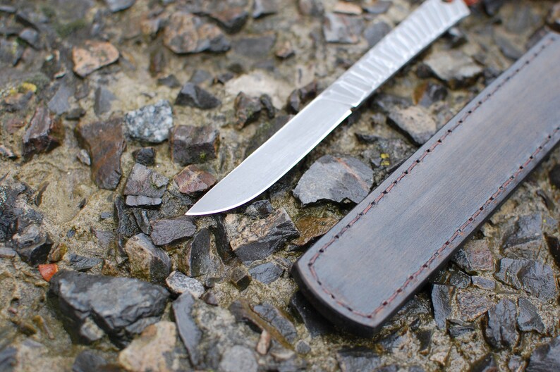 May include: A silver knife with a leather sheath. The knife has a textured blade and the sheath has a stitched edge.
