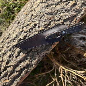 May include: A brown leather knife sheath with a metal snap closure and a leather strap. The sheath is lying on a piece of tree bark.