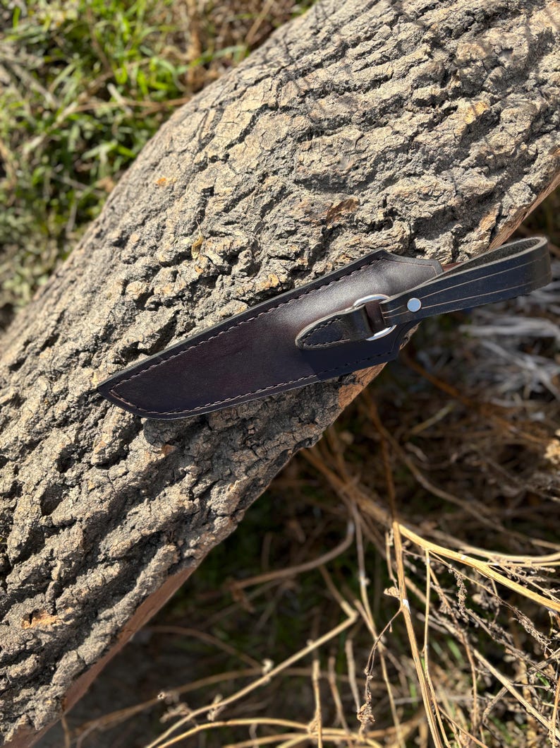 May include: A brown leather knife sheath with a metal snap closure and a leather strap. The sheath is lying on a piece of tree bark.