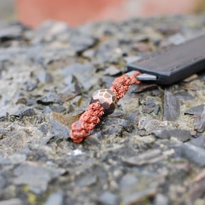 May include: A close-up of a brown leather knife sheath with a red and brown cord and a decorative bead. The bead is a geometric shape with a brown and white pattern.