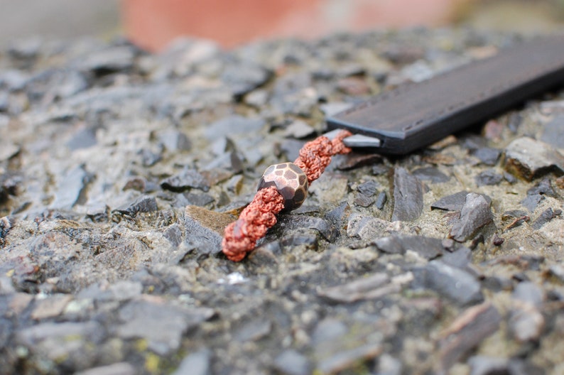 May include: A close-up of a brown leather knife sheath with a red and brown cord and a decorative bead. The bead is a geometric shape with a brown and white pattern.